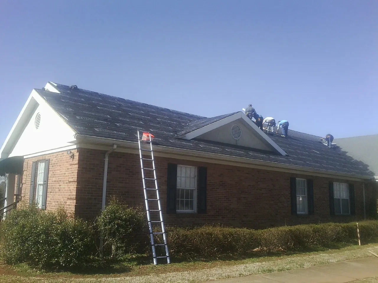 Crew tearing shingles off a brick ranch roof with a ladder set — Emergency Roof Repair work by our crew in Thor