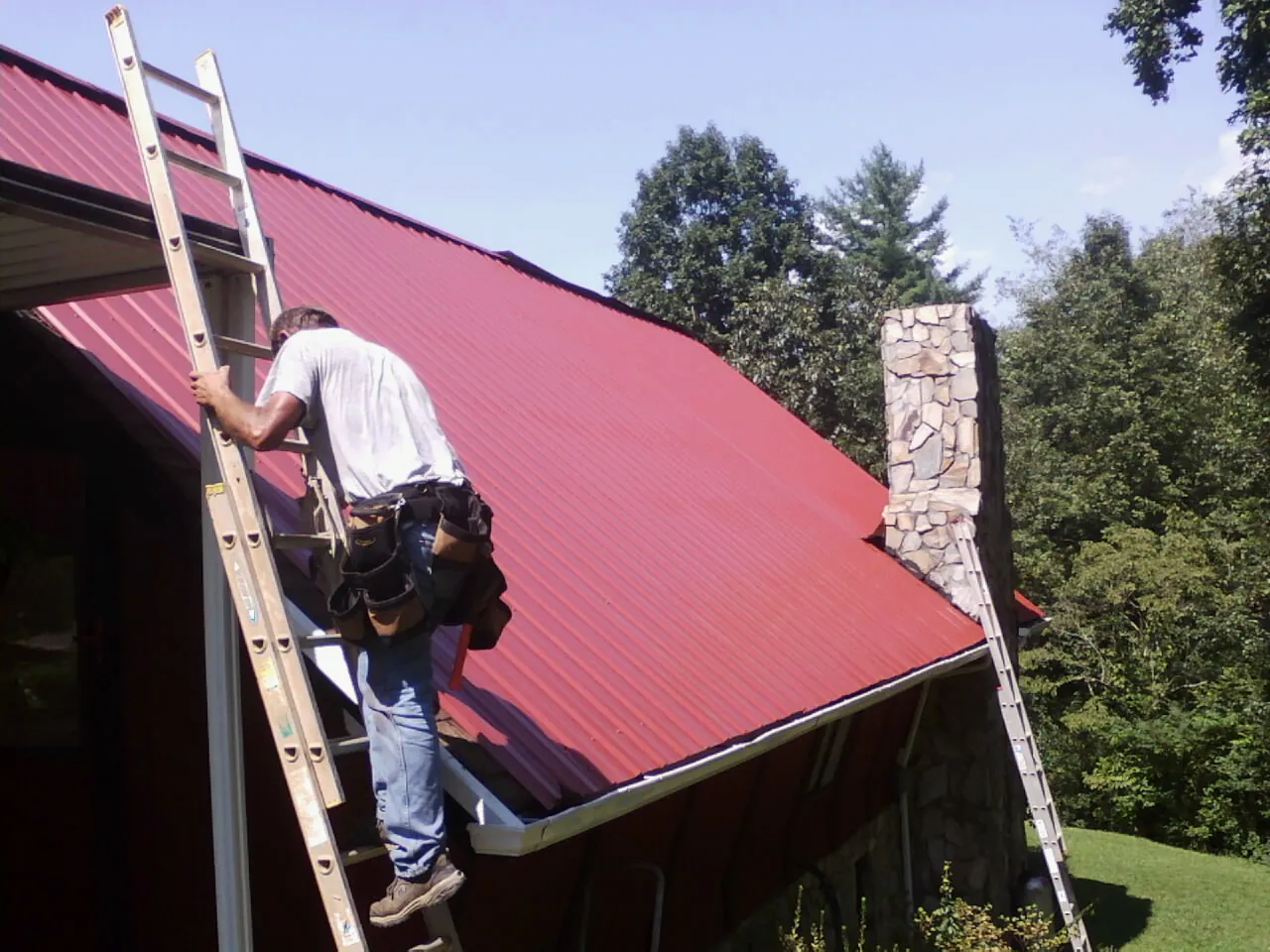 Roofer on a ladder beside a red metal roof and stone chimney — Soffit & Fascia Repair by Roofing Brothers in Thor